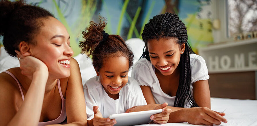 A mum and her two daughters smiling and sharing screentime. All three are looking at the same tablet screen.