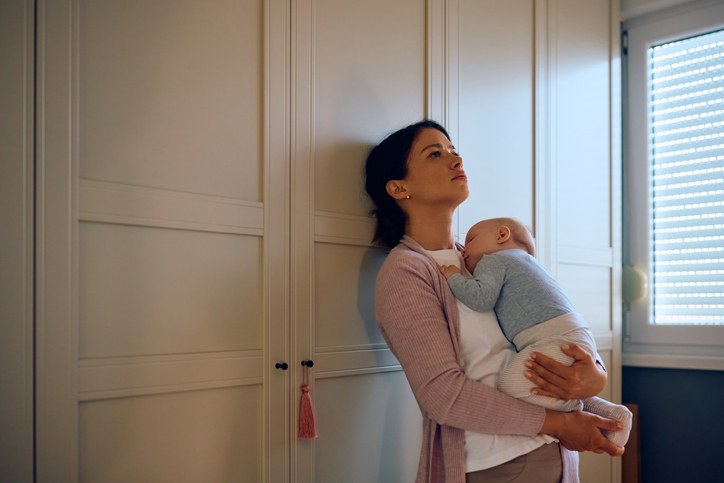 Parent holding a sleeping baby while leaning against a wall and looking upwards